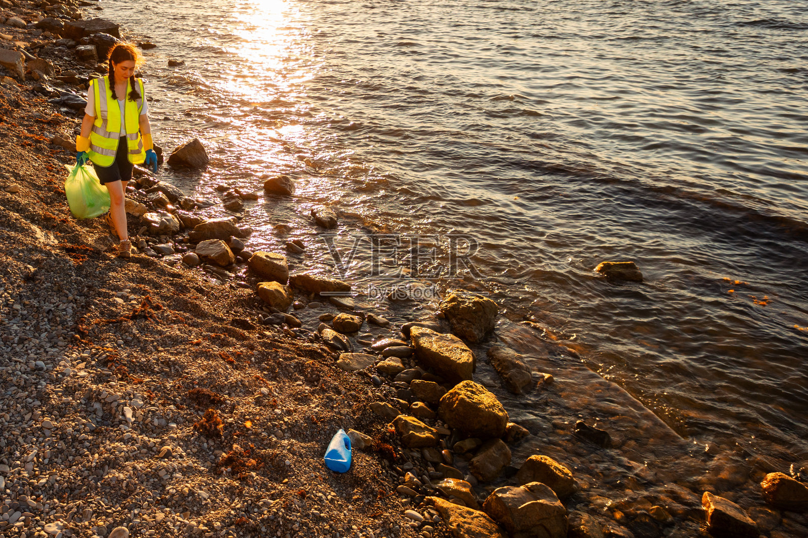 俯视图年轻的白人女性志愿者穿着背心和橡胶手套,拿着装满垃圾的塑料袋沿着海岸走。副本的空间。生态灾害概念照片摄影图片