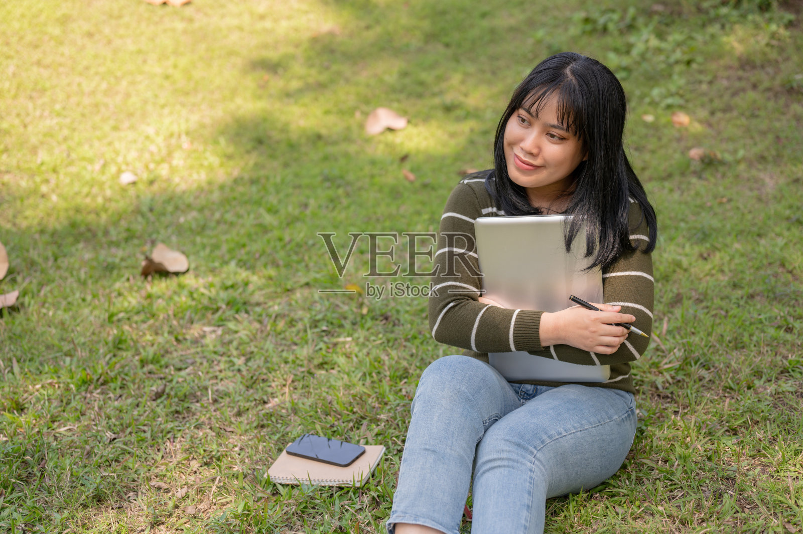 一位乐观的亚洲女大学生拿着笔记本电脑坐在校园公园的草地上。照片摄影图片