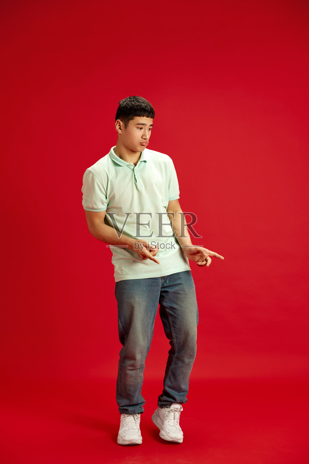 20多岁的亚洲年轻人穿着基本的休闲装,在红色的工作室背景下跳舞,手指指向一边。照片摄影图片