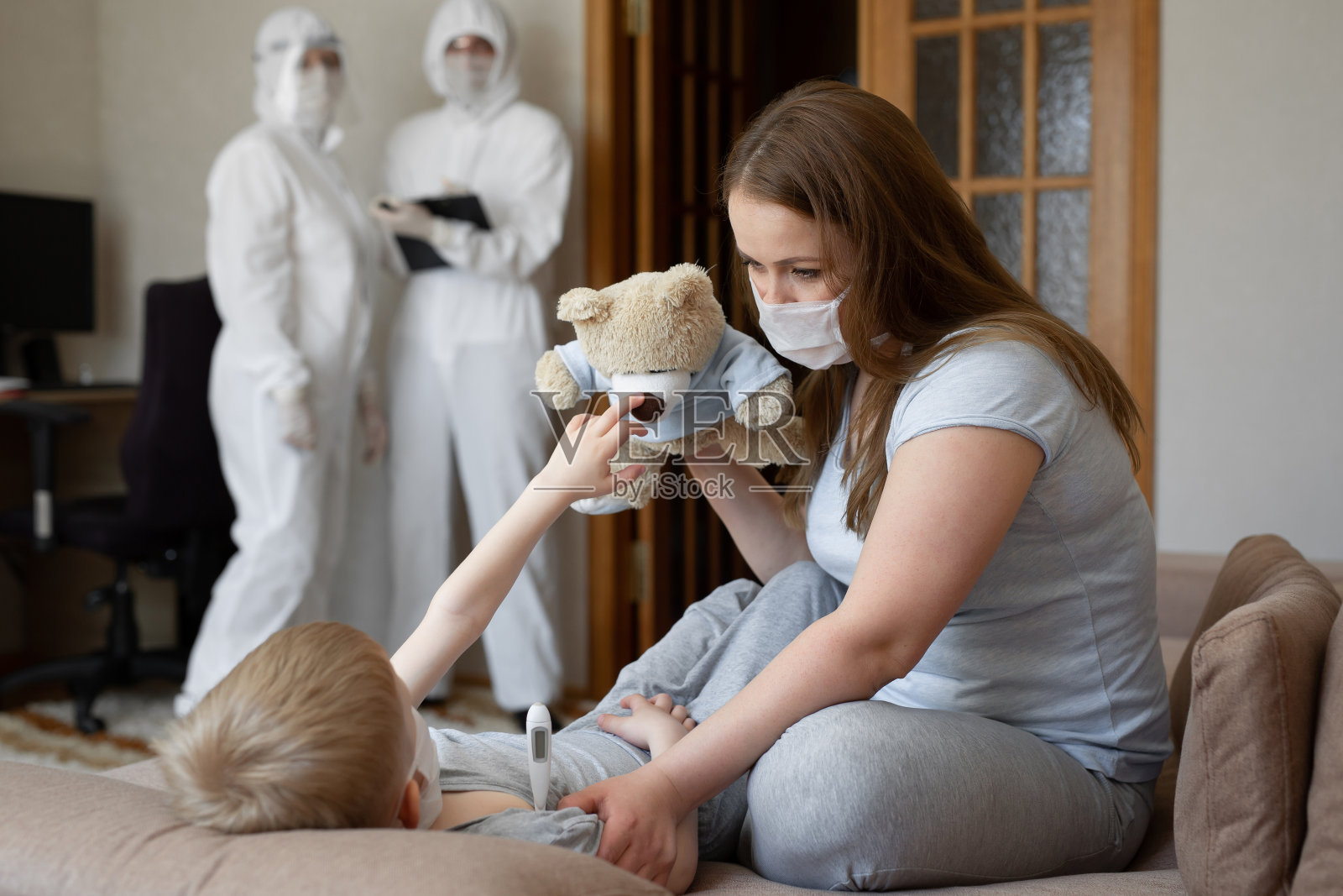 妈妈测量宝宝的体温。穿着防护服的医生在家里照顾病人。冠状病毒,covid-19。照片摄影图片