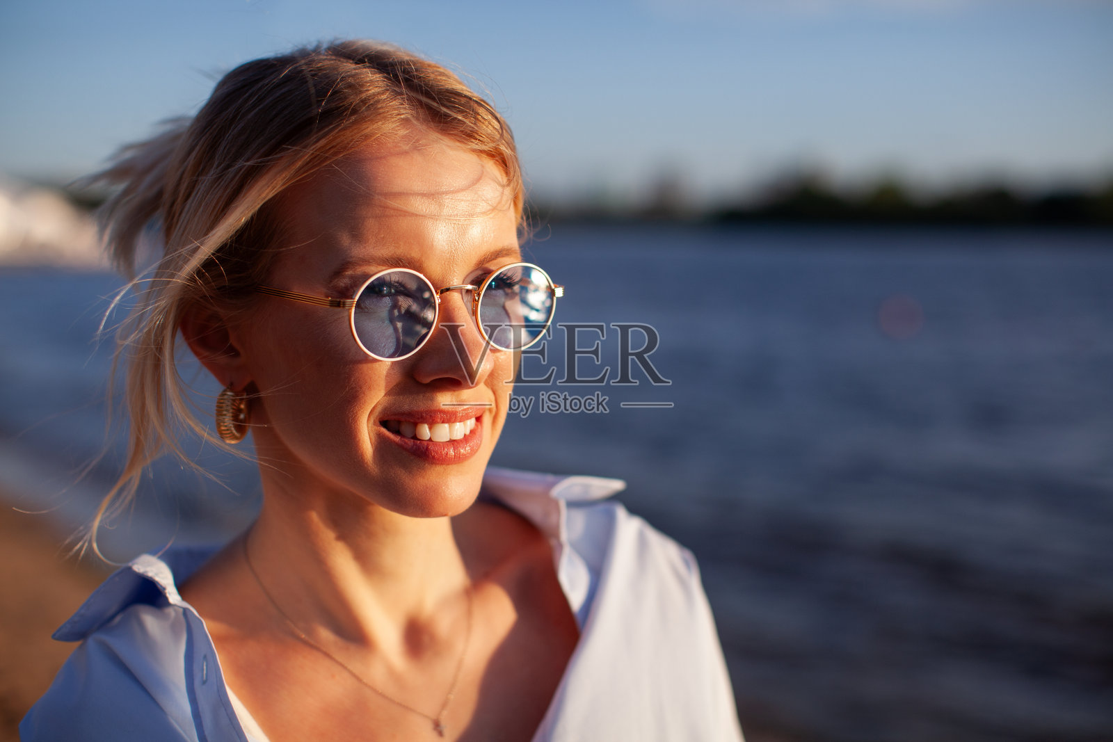 一个美丽的年轻女子戴着太阳镜,在日落的时候微笑着,在水面上反射,享受大自然的闲暇时光照片摄影图片