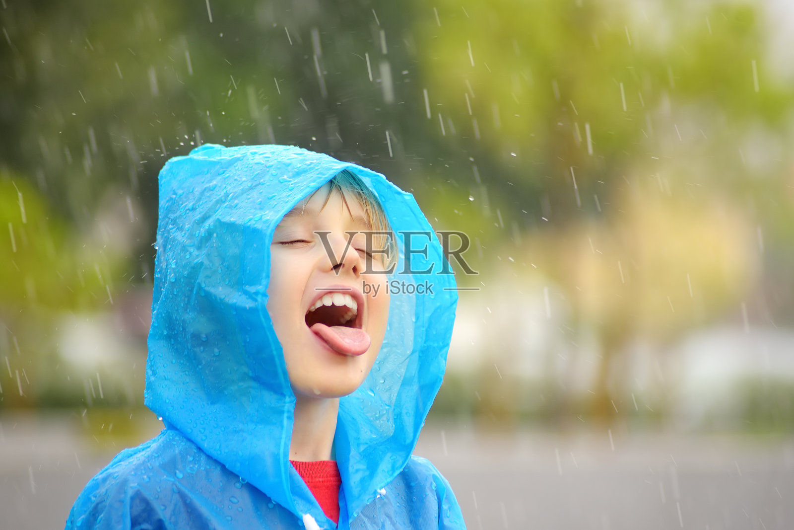 倾盆大雨中一个快乐的孩子的肖像。一个穿着蓝色雨衣的快乐男孩在倾盆大雨中走着,舌头上沾着水滴。照片摄影图片
