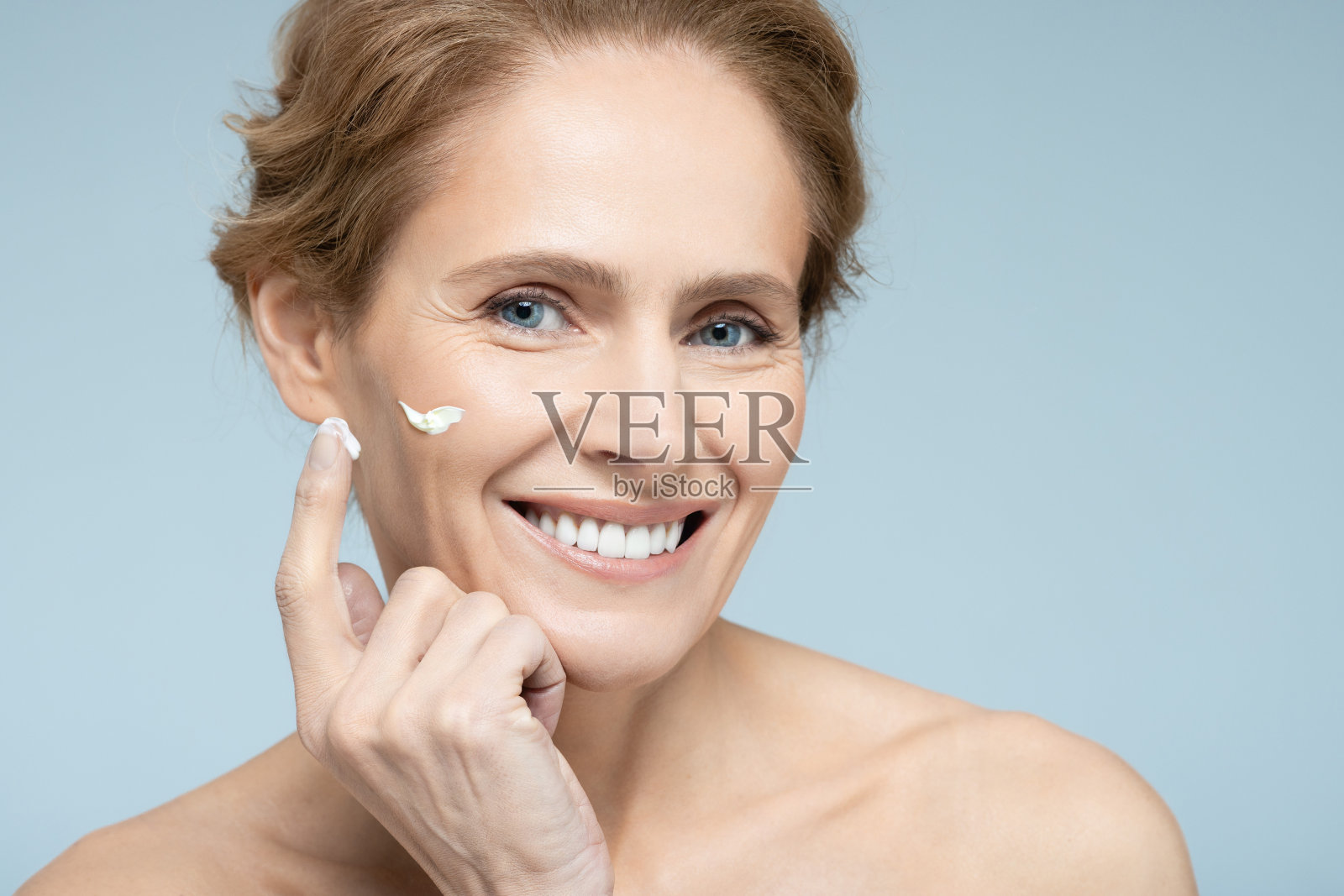 中年妇女在脸上涂抹面霜和抗衰老化妆品,蓝色背景。女性护肤用抗衰老化妆品照片摄影图片