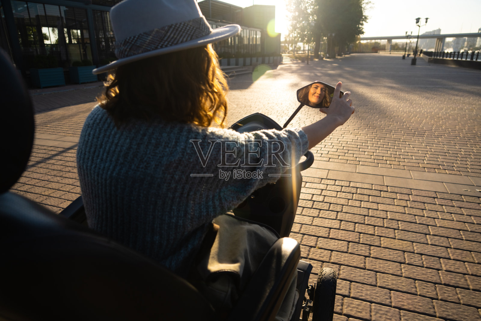 戴着灰色帽子和毛衣的女游客骑着四轮电动滑板车走在城市街道上。夕阳背光。照片摄影图片