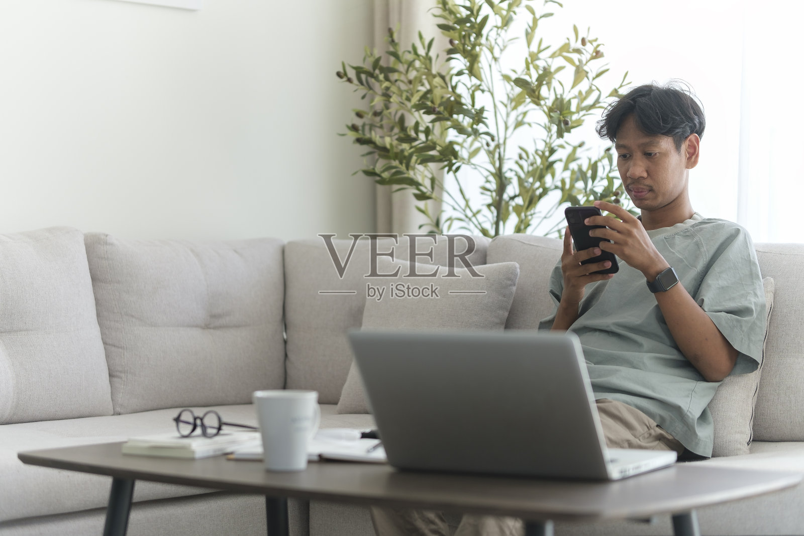 微笑的亚洲男性的肖像,用智能手机坐在地板上,用笔记本电脑,看短信,靠在沙发上,在家庭办公室工作。照片摄影图片