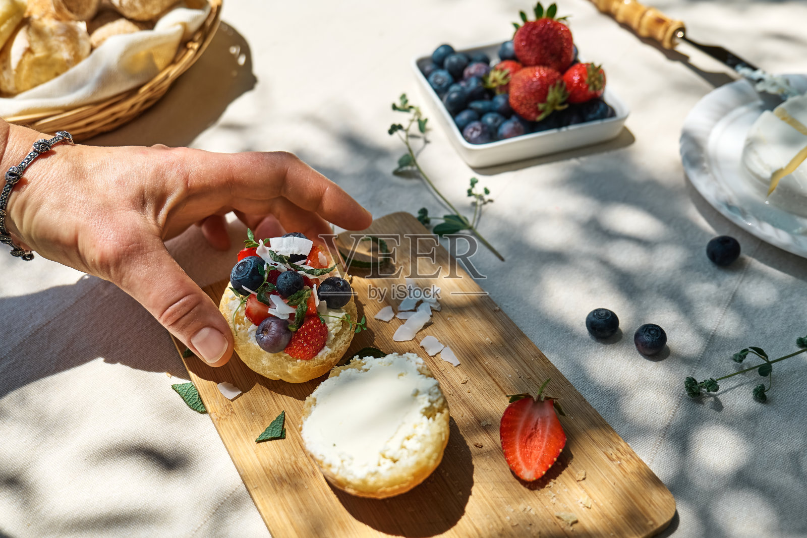 一位不知名的女士在花园里为健康早餐制作甜美的迷你三明治。烤面包配奶油芝士、蓝莓、草莓、椰子片和香草,适合夏季野餐或早午餐照片摄影图片