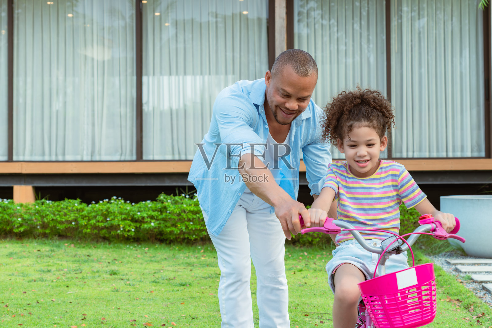 一个非洲父亲在闲暇时间指导他的卷发的小学年龄的女儿周末在院子里骑粉红色的自行车锻炼。照片摄影图片