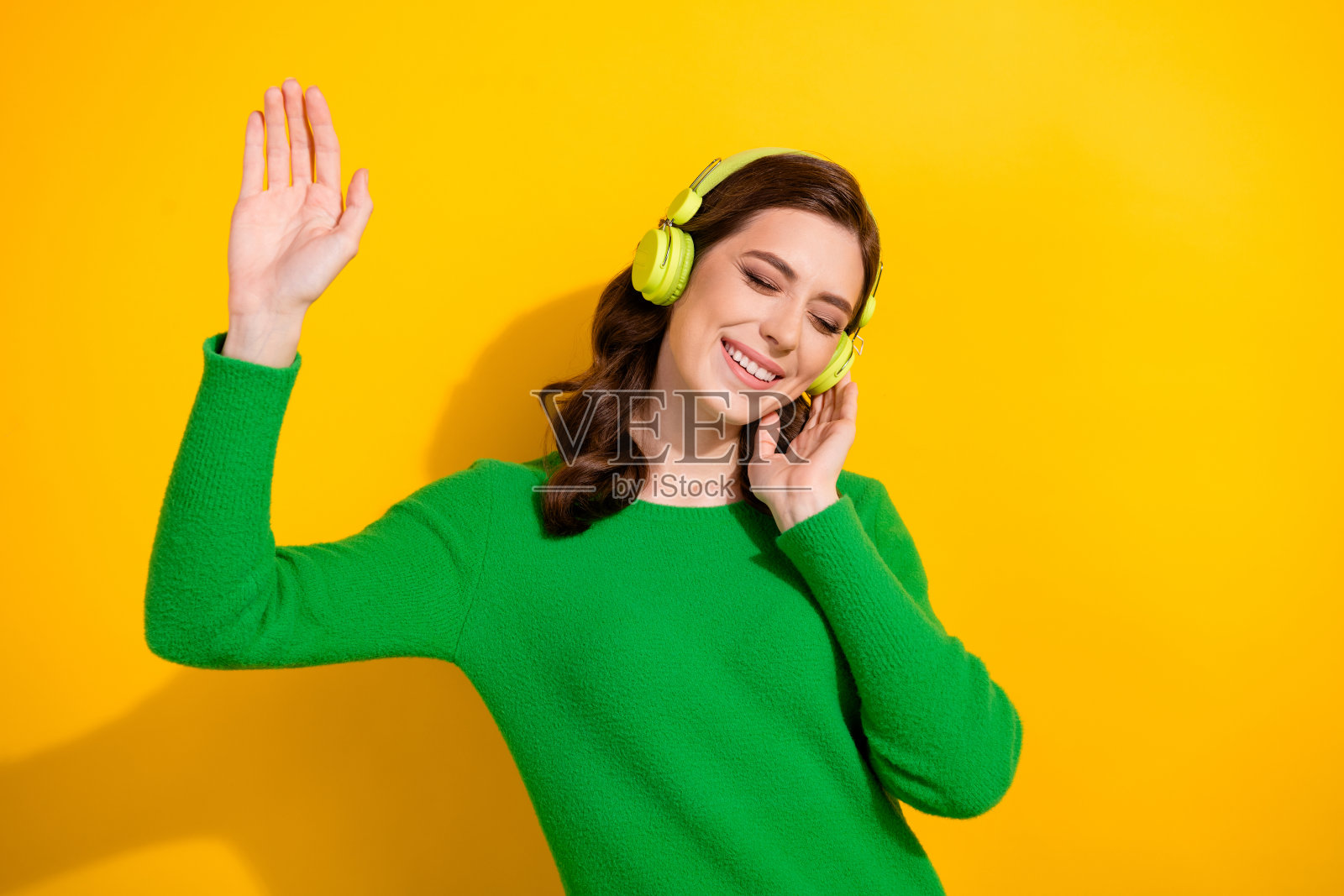 照片中,年轻漂亮的女士穿着绿色的套头衫,举起手掌,心情愉快地跳舞,触摸耳机,孤立在黄色背景上照片摄影图片