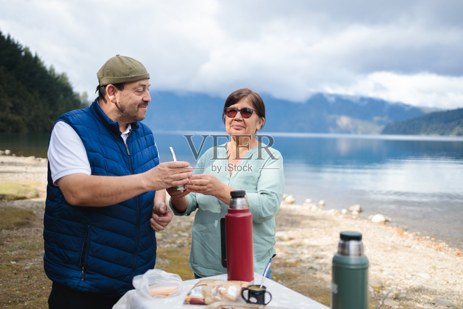 拉丁中年夫妇在美丽的巴塔哥尼亚山湖边户外喝马黛茶。照片摄影图片