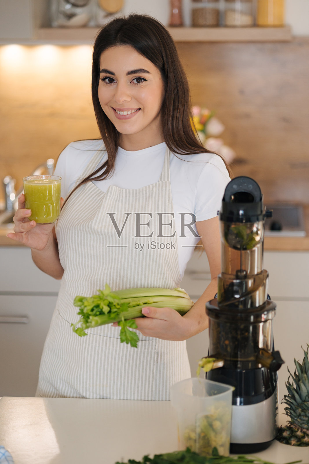 穿着围裙站在厨房里的美丽而微笑的女性画像。女子一手拿着绿色健康饮料,一手拿着芹菜。排毒饮食。自制的果汁。素食者照片摄影图片