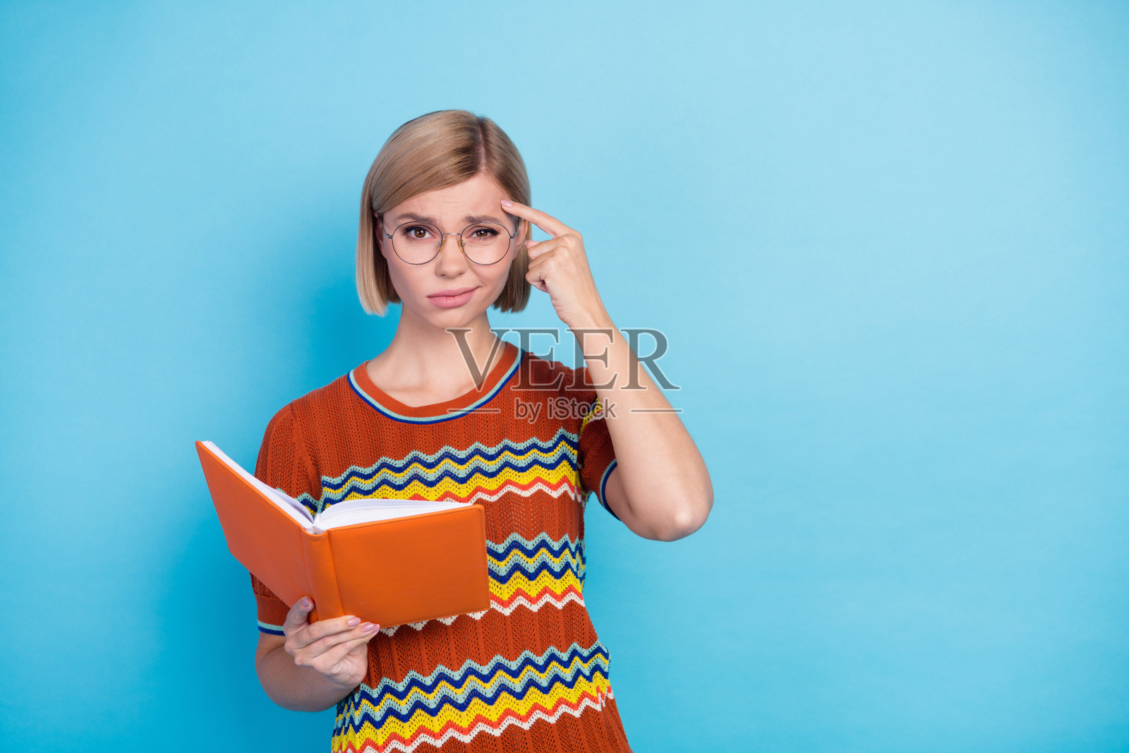 照片中的压力妇女穿着针织t恤,戴着眼镜,拿着书,手指在头上,准备考试孤立的蓝色背景照片摄影图片