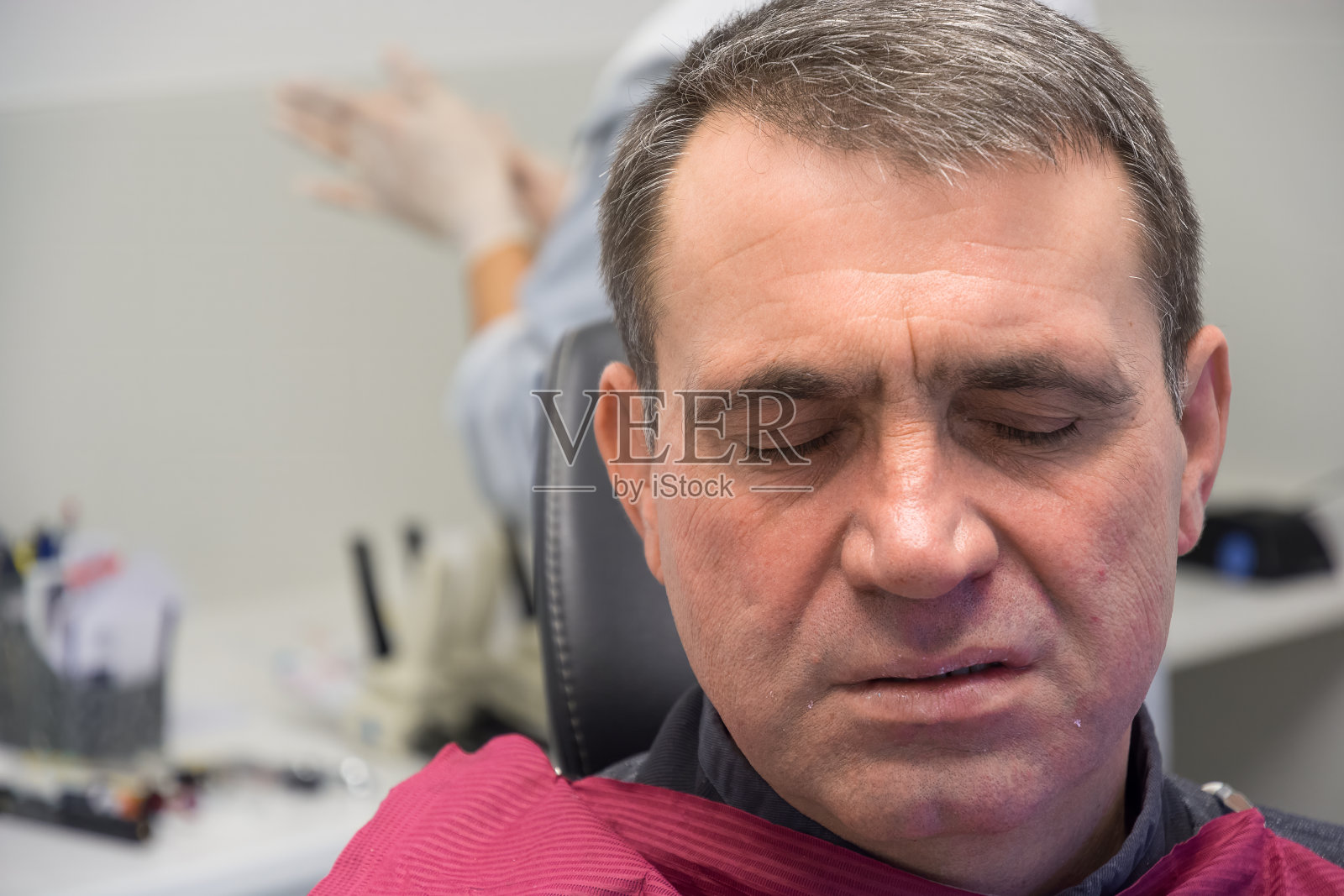 一个脸因疼痛而扭曲的男人在看牙医时闭上了眼睛。在模糊的背景中可以看到医生戴着医用手套的手。牙科治疗无痛的概念。照片摄影图片