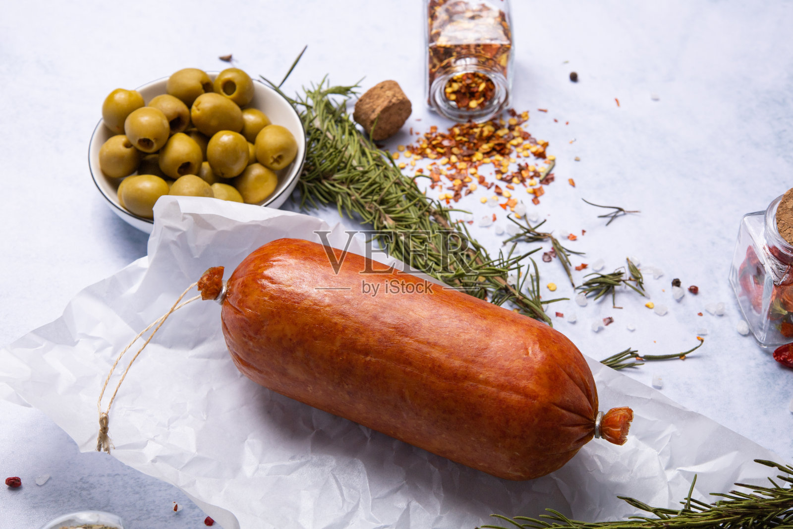 在蓝色的桌面上,熏香肠被香料和香草包围着,旁边是橄榄和芥末照片摄影图片