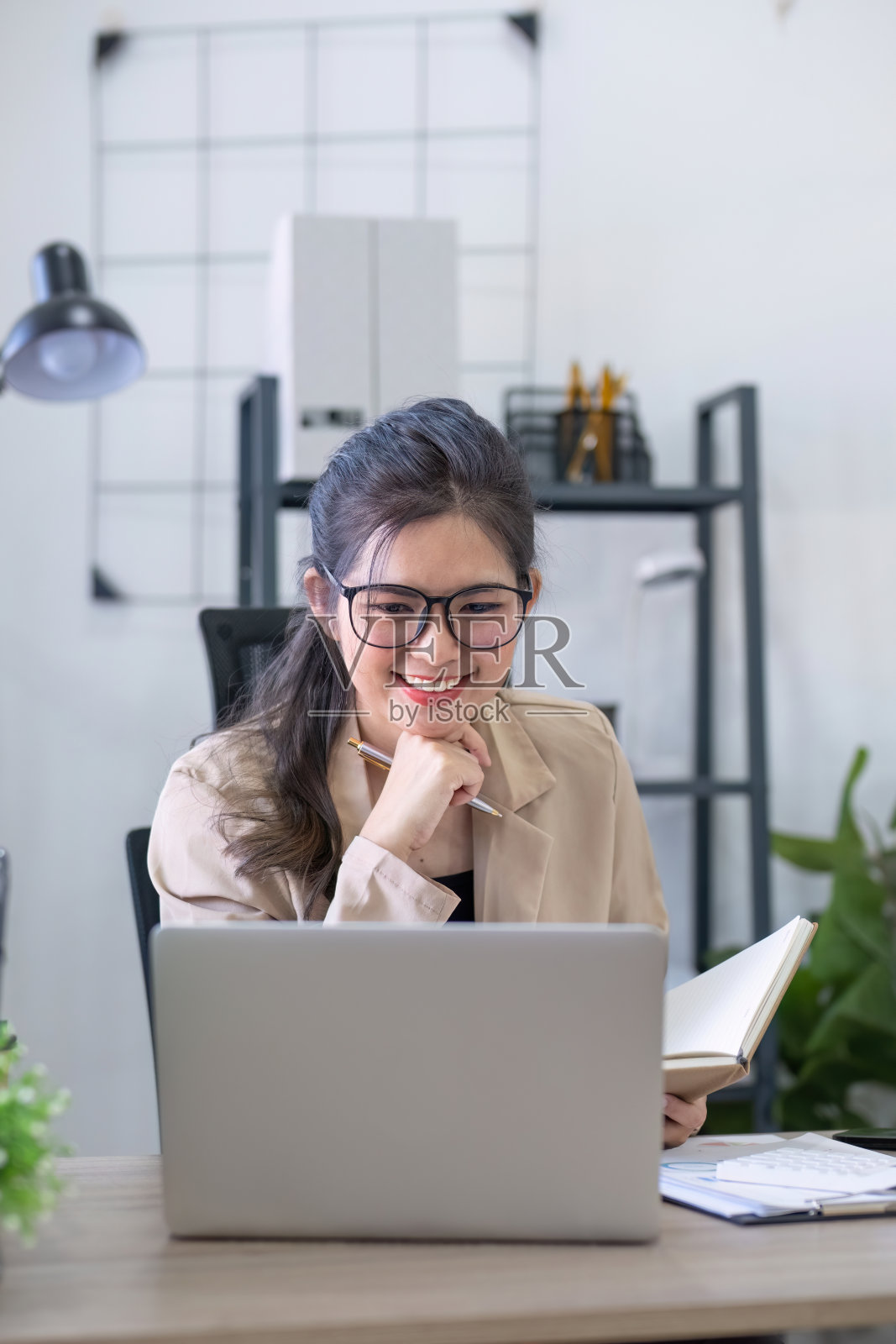 亚洲女商人或会计愉快地坐着,用笔记本电脑工作在金融和企业管理。在办公室的桌子上装饰着绿色的植物。照片摄影图片