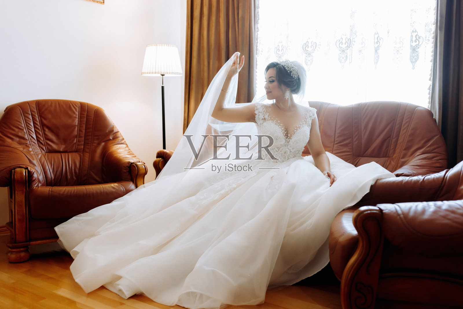 美丽的新娘穿着白色的礼服,漂亮的头发和化妆,坐在豪华的沙发上。盛装打扮,准备婚礼照片摄影图片