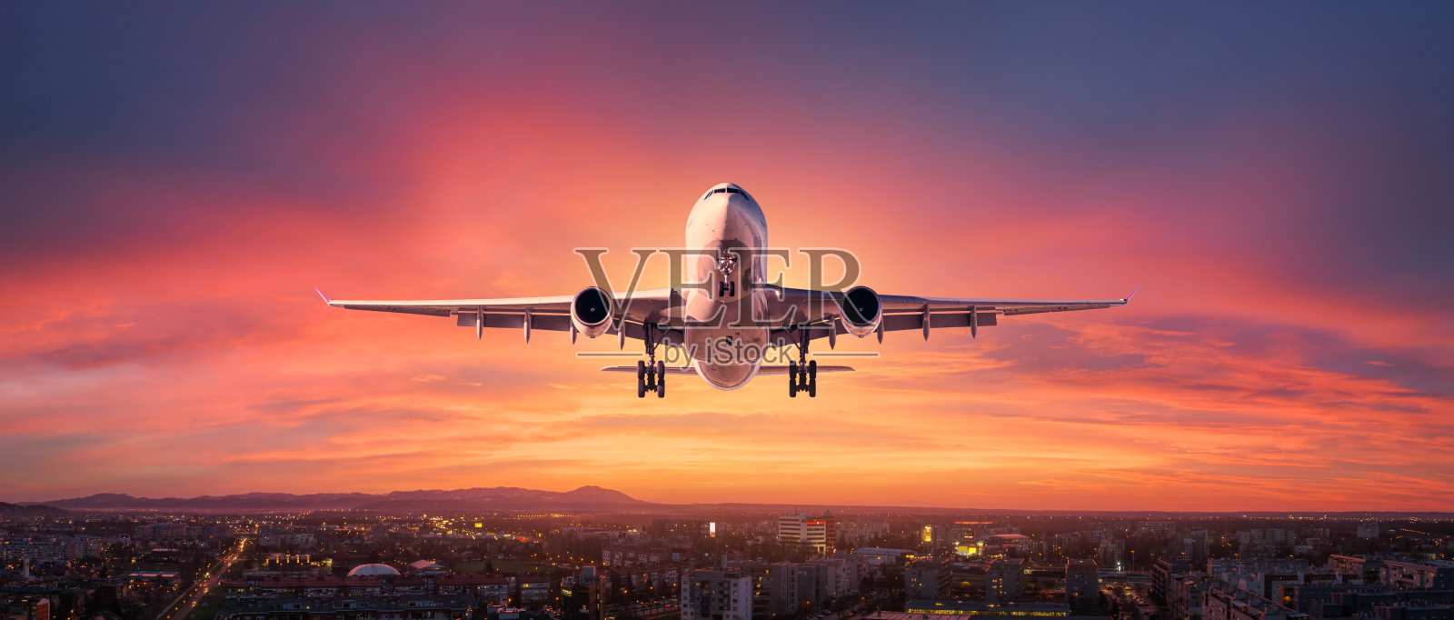 夕阳下,一架飞机在城市上空五颜六色的天空中飞行。有客机的风景,天际线,黄昏时紫色的天空和红色和粉红色的云。飞机将在黄昏降落。飞机鸟瞰图照片摄影图片