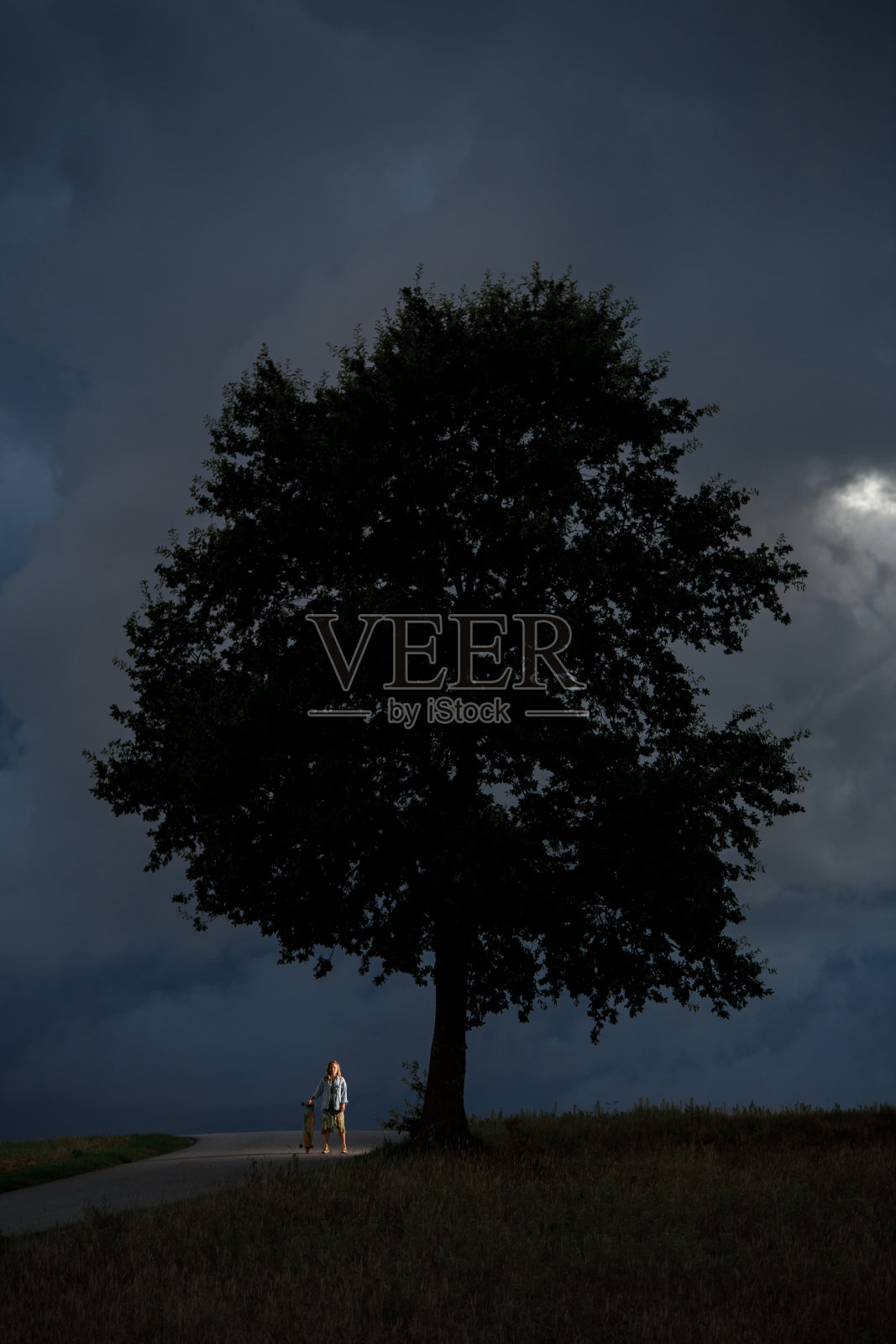 一个小女孩站在路边,手里拿着她的长板,旁边只有一棵树照片摄影图片