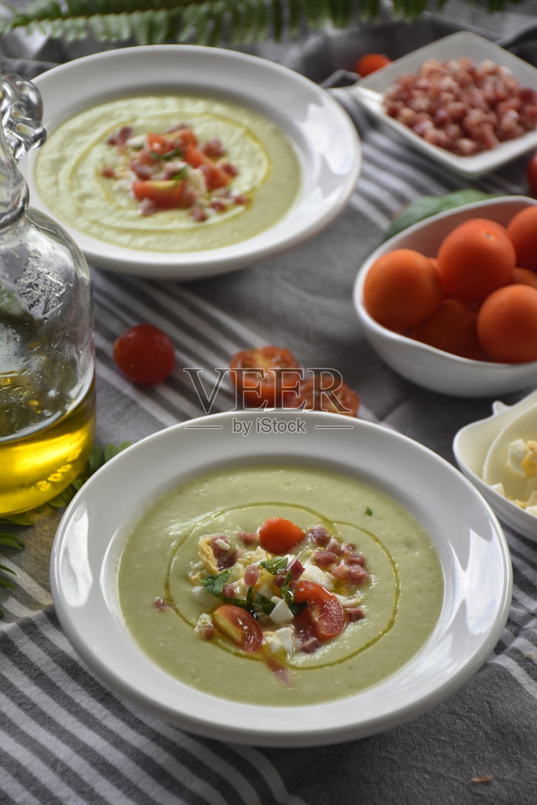 凉拌牛油果西班牙凉菜汤,配以西红柿、煮熟的鸡蛋和火腿,放在一个白色的碗里,桌子上放着一条灰色的厨房毛巾、橄榄油和香料植物。照片摄影图片