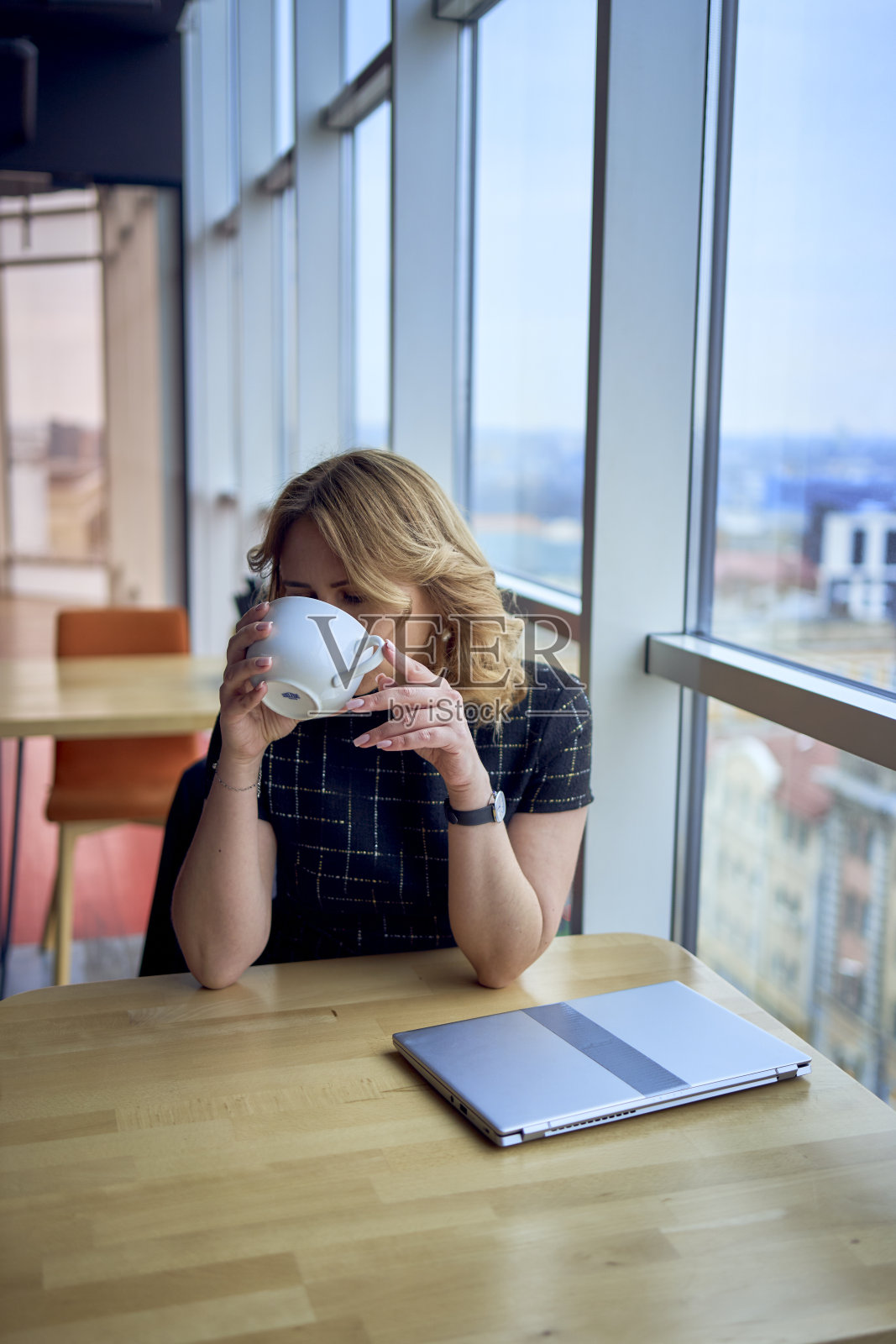 迷人的成熟女人坐在咖啡馆的桌子上喝咖啡,旁边是全景窗户,可以看到城市的景色照片摄影图片