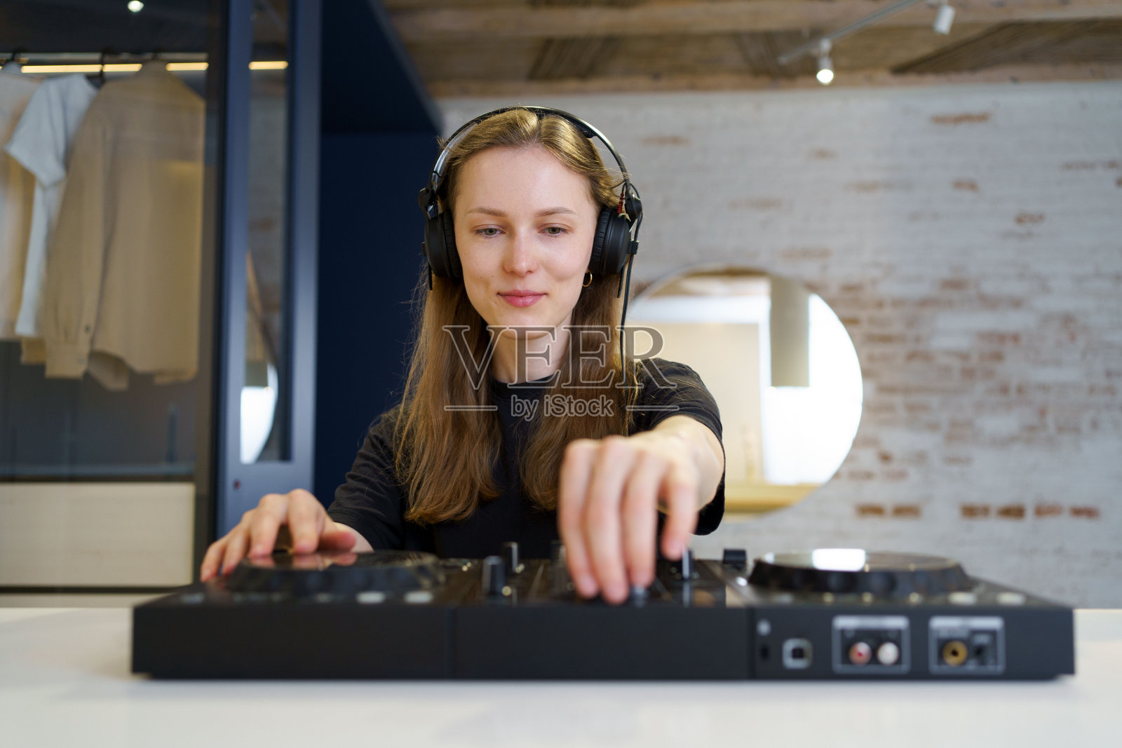 年轻的女DJ穿着黑色衣服,戴着耳机,在家里的房间里用控制器混合音乐。DJ台的概念,表演和休闲时间照片摄影图片