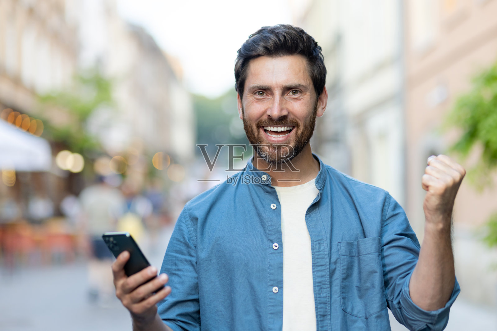 一个穿着蓝色衬衫站在外面街道上的快乐年轻人的肖像,他手里拿着手机,看着镜头,用手做出胜利的手势照片摄影图片