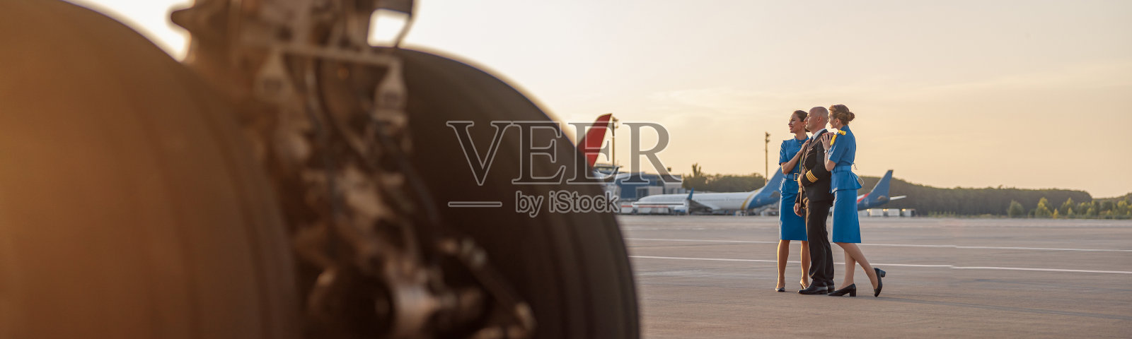 日落时分,一名男飞行员与两名身穿蓝色制服的空姐站在机场候机楼,摆姿势拍照照片摄影图片