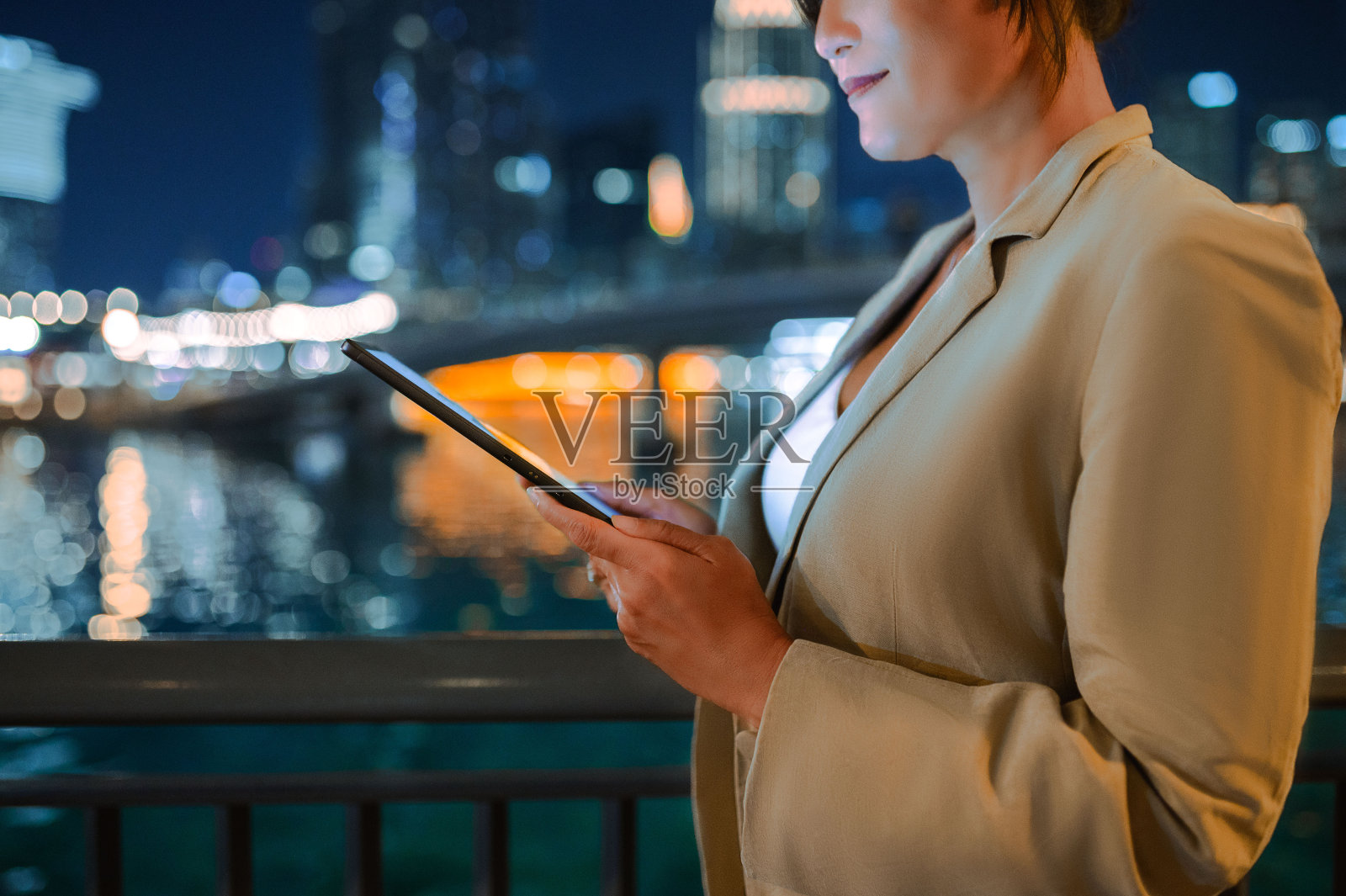裁剪的女商人双手拿着数码平板电脑,背景是城市里被照亮的河流和建筑物照片摄影图片