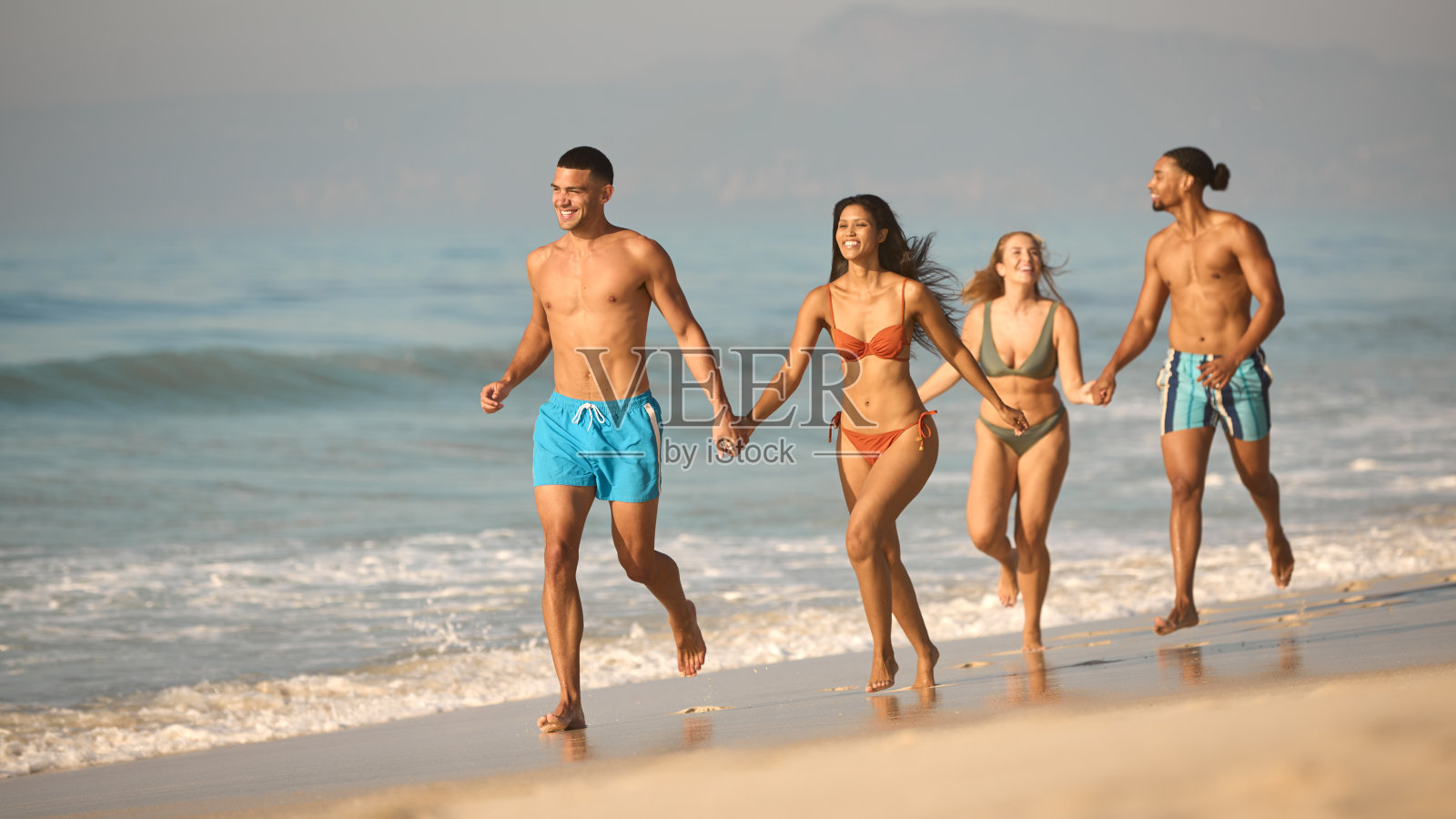 一对夫妇和朋友在度假时穿着泳装,手牵着手沿着海滩岸线奔跑照片摄影图片