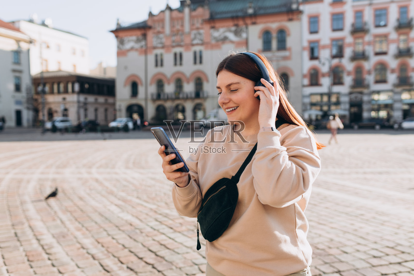 一个可爱的年轻女孩在城市背景下通过无线耳机听音乐的特写。音乐爱好者享受音乐。妇女用手提电话录下声音照片摄影图片