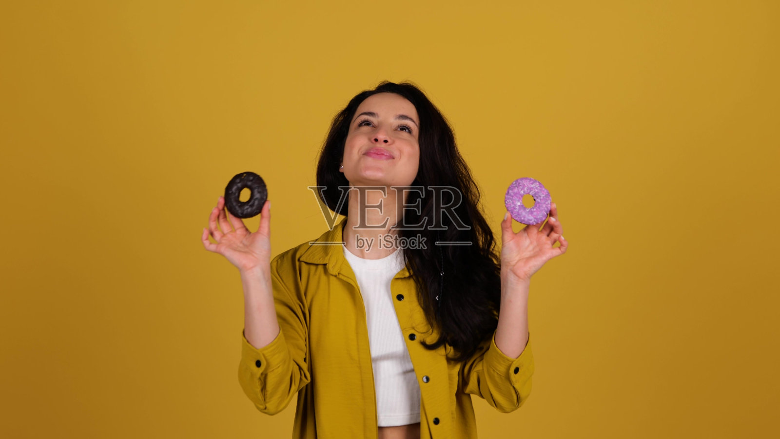 年轻漂亮的女人拿着甜甜圈靠近脸,同时在工作室里摆姿势,手里拿着美味的糖果。人们生活方式的饮食观念。拷贝空间。实时照片摄影图片