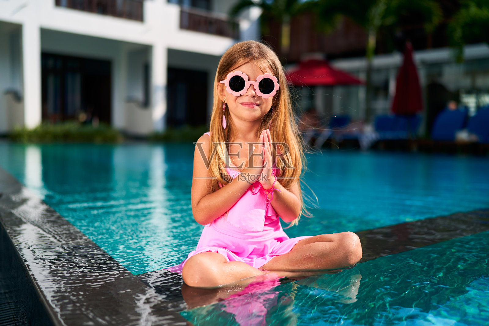 小女孩穿着粉红色的裙子,戴着墨镜,在泳池边摆出莲花的姿势。孩子练习瑜伽,合十礼,享受热带假期。健康,家庭度假概念与孩子放松。照片摄影图片