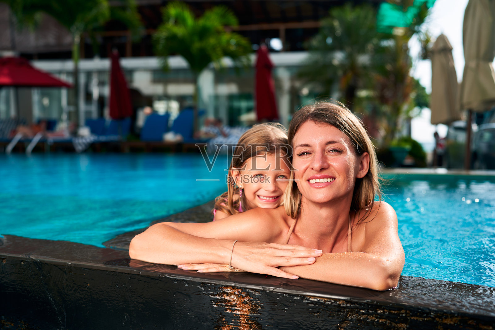 微笑的母亲,女儿享受泳池边拥抱在热带度假胜地。孩子,女人拥抱,在阳光明媚的碧水边欢笑。全家放松,分享暑假的爱。无忧无虑的时刻,温暖的bond池。照片摄影图片