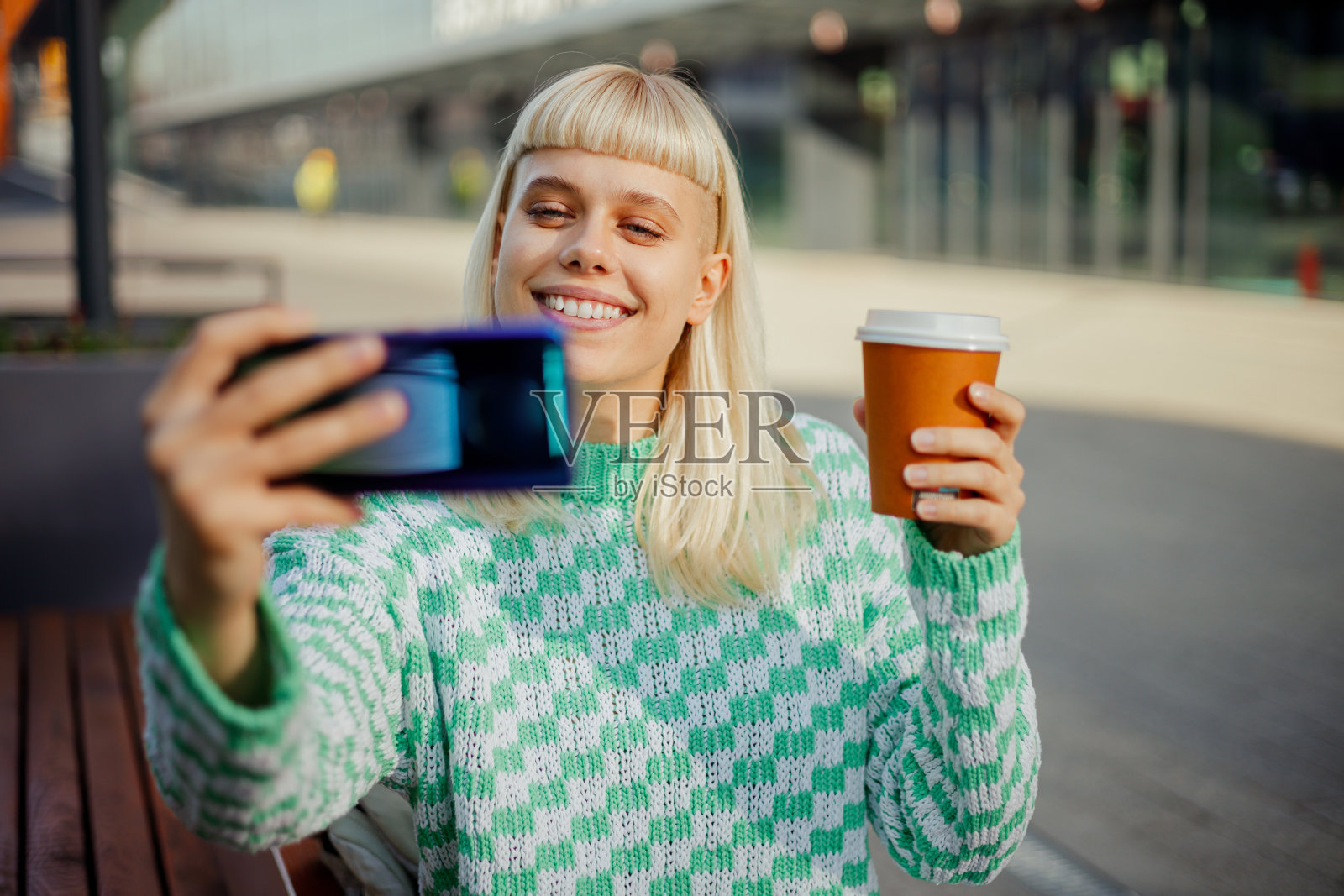 一个都市女孩坐在街上,端着咖啡自拍。照片摄影图片