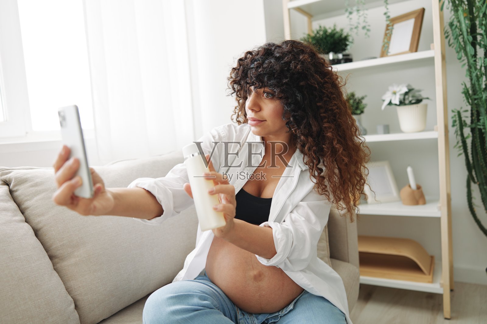 孕妇微笑博主展示婴儿奶瓶拍摄自己的手机,视频聊天坐在家里的沙发上,在怀孕的最后一个月的自由职业者照片摄影图片