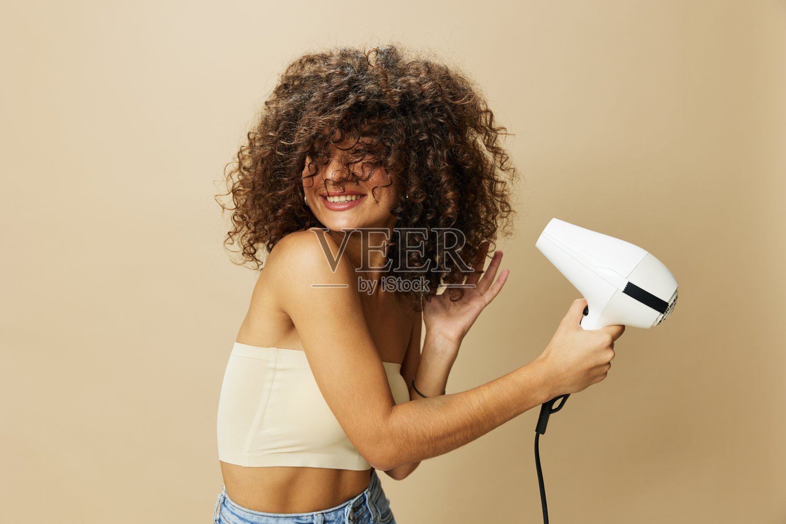 女人用吹风机吹干卷曲的非洲发型,家用美容护理定型产品,米色背景上的微笑照片摄影图片