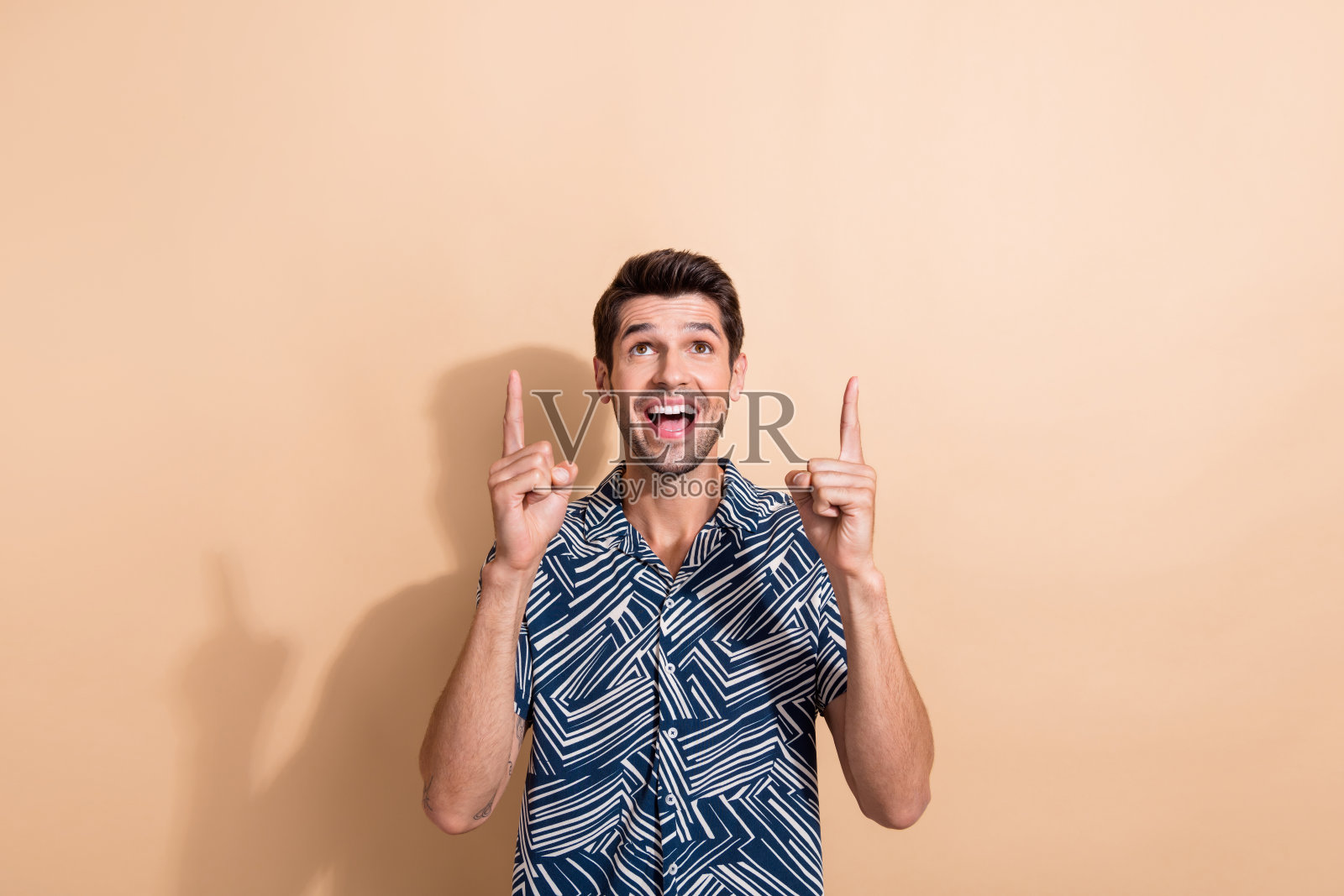 一个有着时髦发型的喜出望外的家伙的肖像,穿着时髦的衬衫,在米色背景上显示着抬头看销售的空白空间照片摄影图片