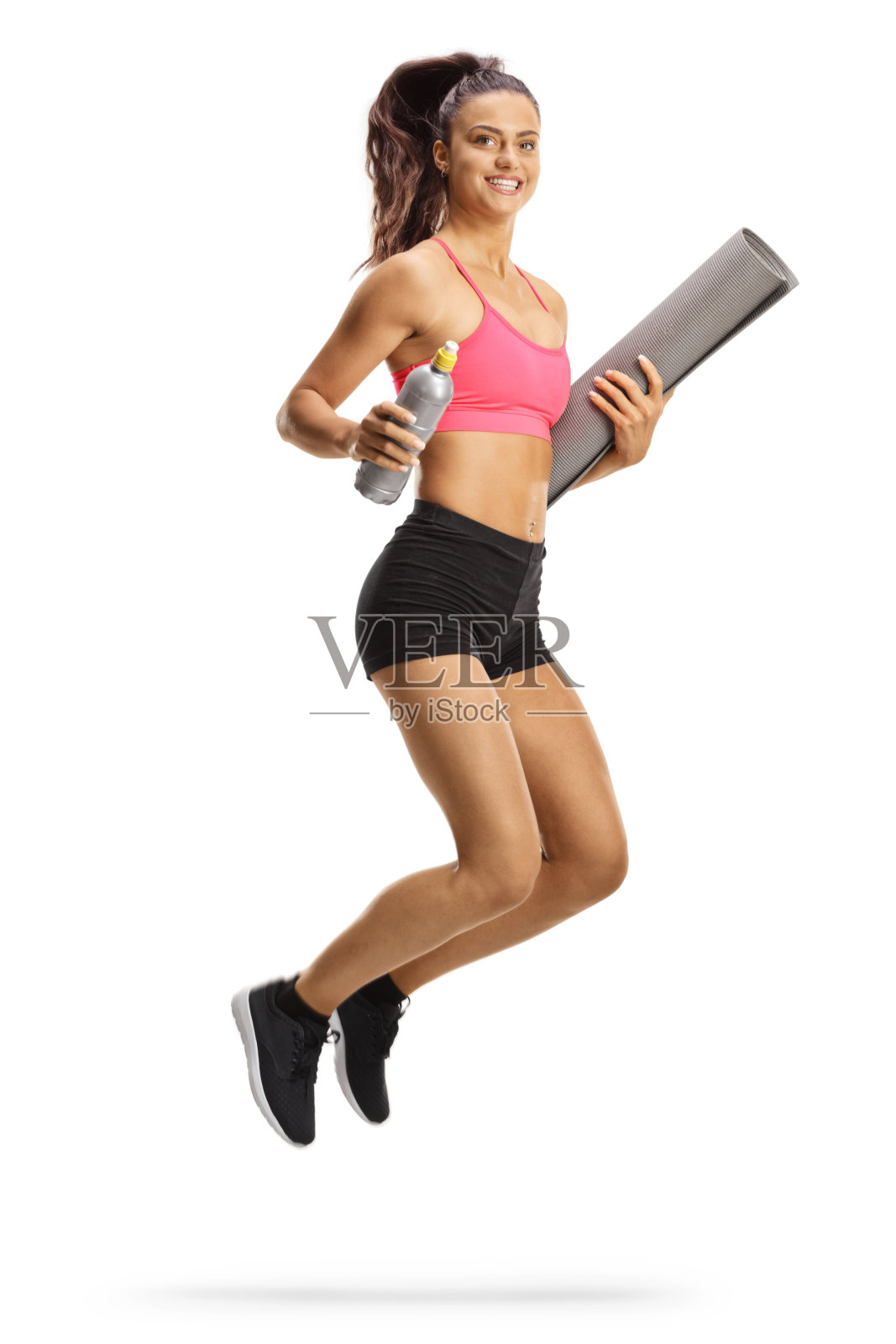 穿着露脐上衣和短裤的年轻女子拿着运动垫和瓶子蹦蹦跳跳照片摄影图片