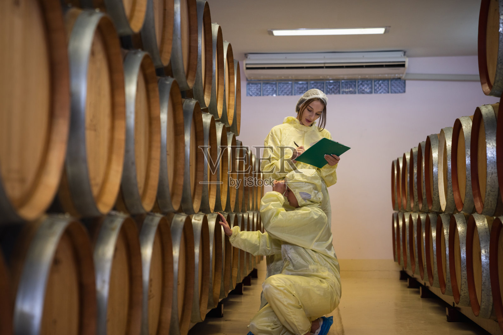 工人们在酒厂检查酒桶。一个女人在一个有木桶的酒窖里品尝和闻葡萄酒。照片摄影图片