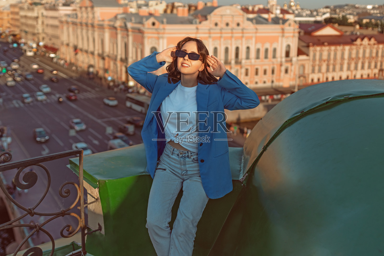 穿着牛仔裤和蓝色夹克的年轻快乐的女人坐在圣彼得堡的屋顶上休息照片摄影图片