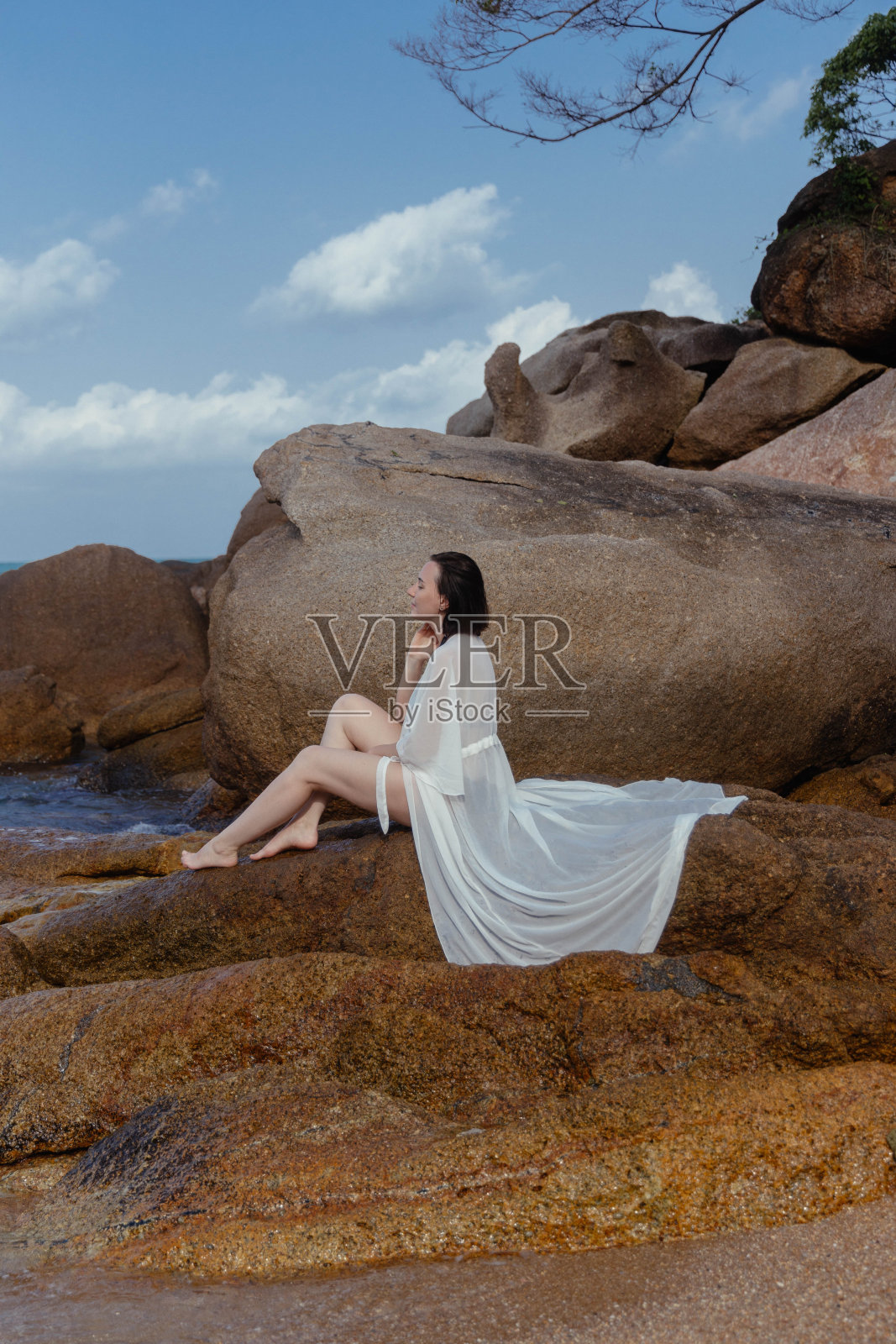一名身穿飘逸白色连衣裙的女子在海边的岩石上享受宁静的时刻,海水轻轻地拍打着她照片摄影图片