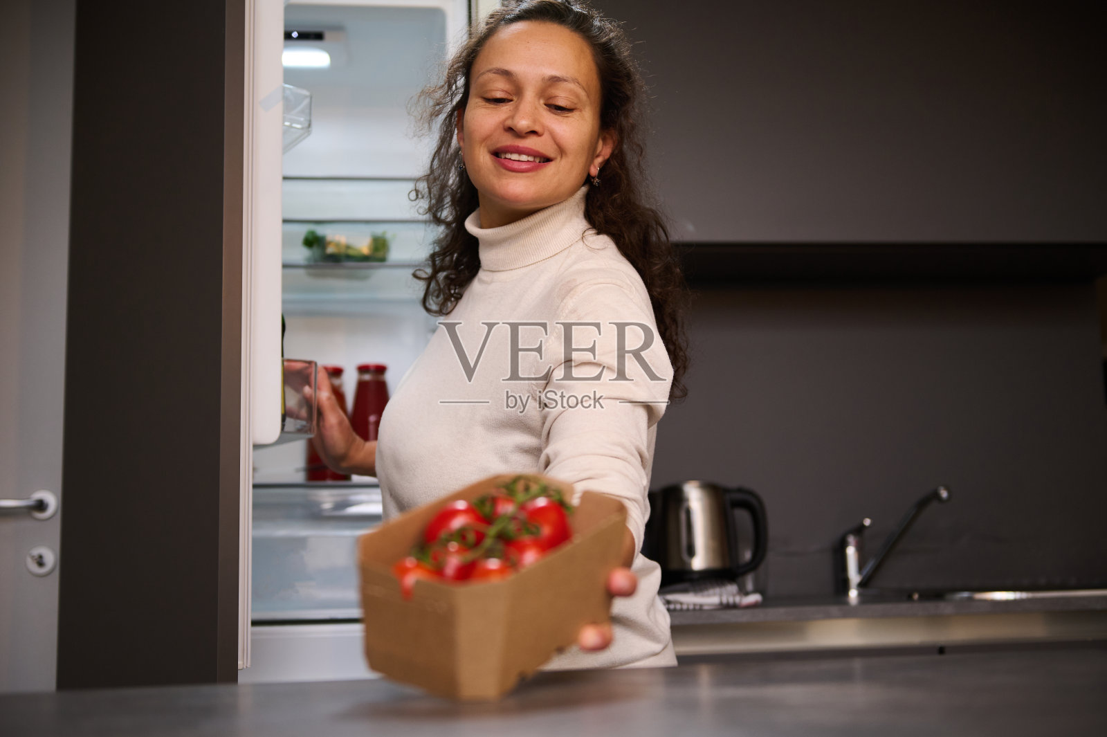 微笑的女人在桌子上放着装有健康有机西红柿樱桃的纸板箱,站在家庭厨房的冰箱旁。人。健康的饮食习惯。食品和饮料消费主义照片摄影图片