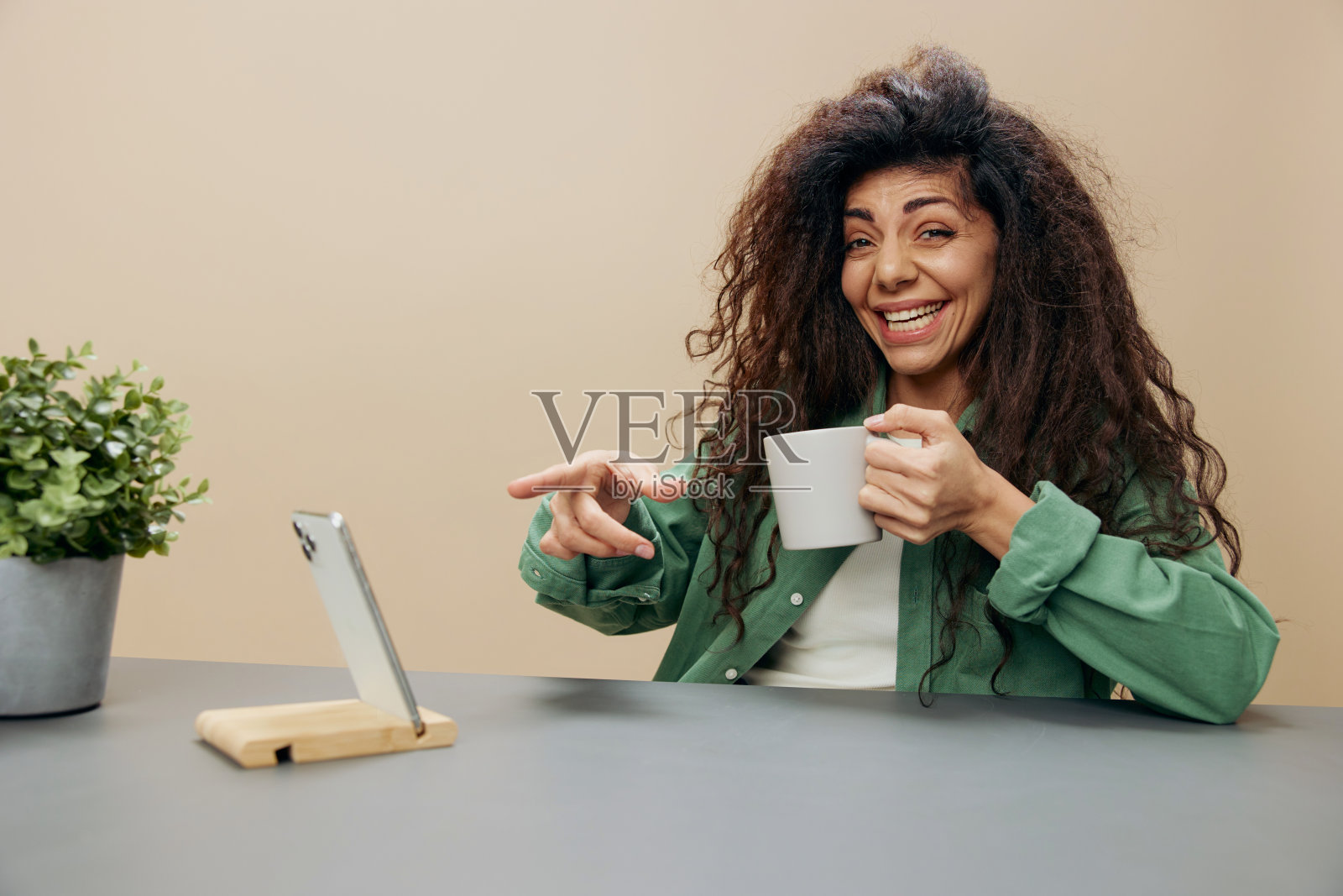 喜出望外的笑着幸福的棕褐色卷曲的拉丁女士拿着一杯茶咖啡用手指着手机屏幕坐在桌子旁,周围是柔和的米色背景。复制空间横幅。概念放松时间照片摄影图片