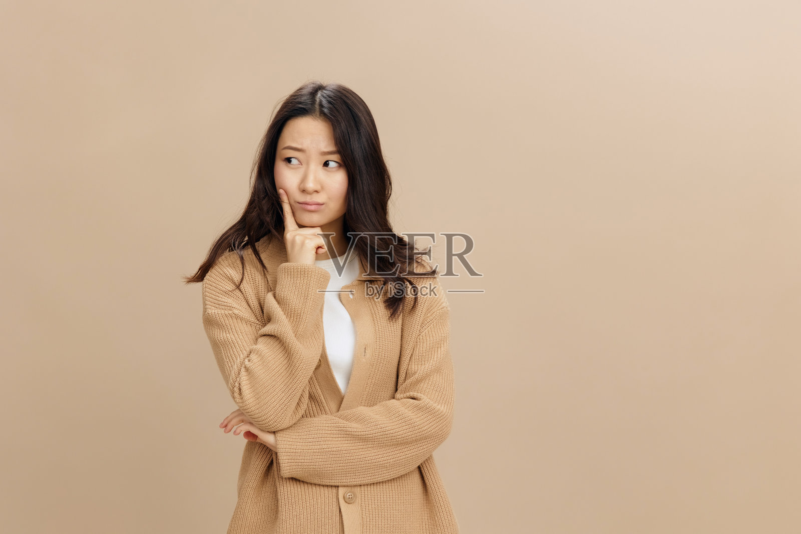 沮丧,沮丧,沉思可爱的亚洲学生年轻女子,穿着温暖的毛衣,手指着脸颊,在米色柔和的工作室背景上独自摆姿势。很酷的时尚报价。时尚概念照片摄影图片