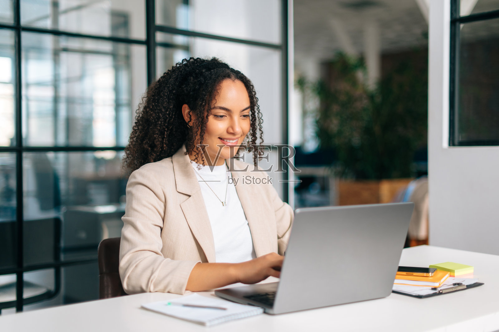 成功的卷发西班牙裔或巴西女性,穿着时尚优雅的衣服,办公室职员,秘书或人力资源经理,坐在她的工作场所,微笑着使用笔记本电脑照片摄影图片