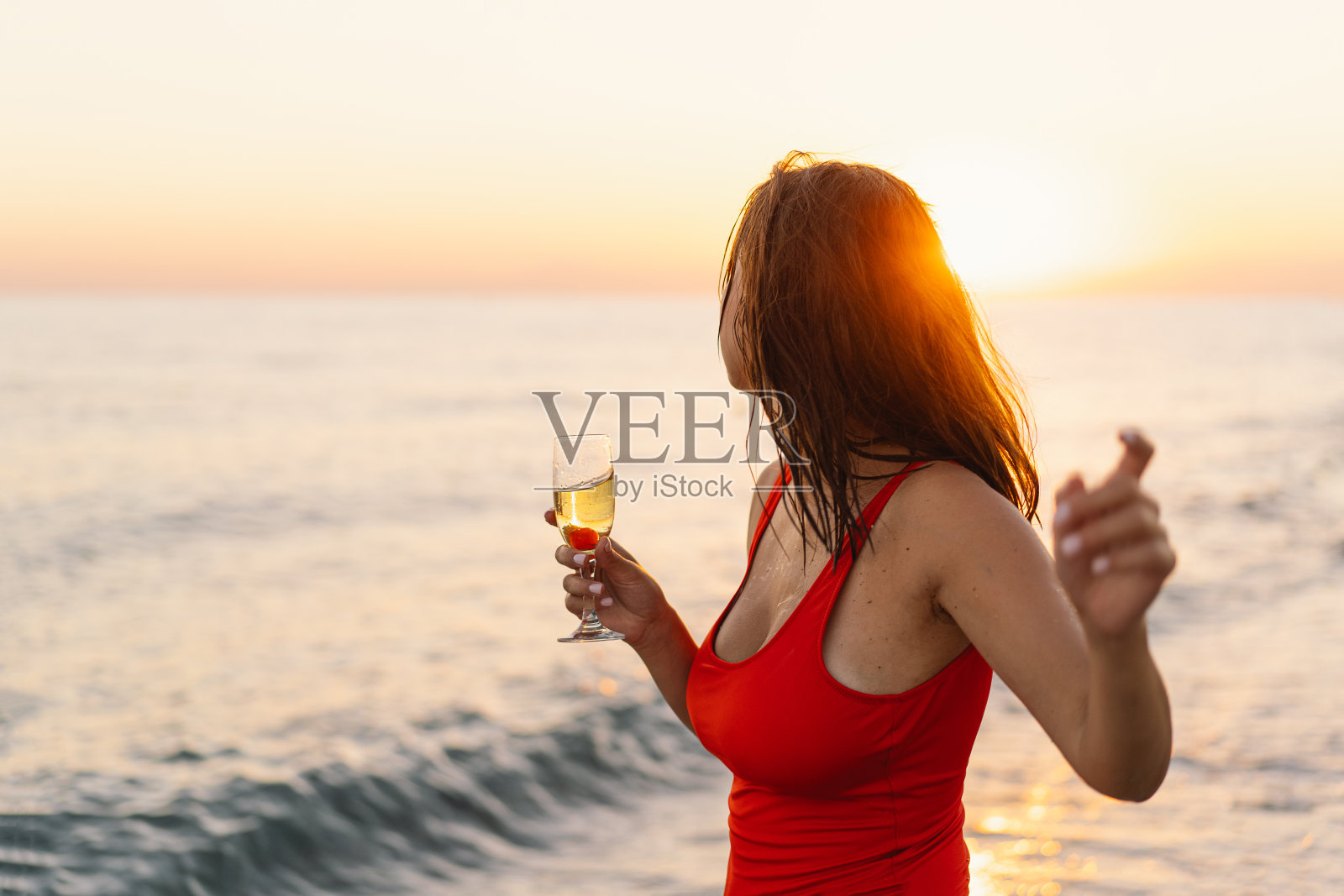 一个穿着鲜艳的红色泳衣的兴高采烈的女人站在海边,手里拿着一杯白葡萄酒。照片摄影图片