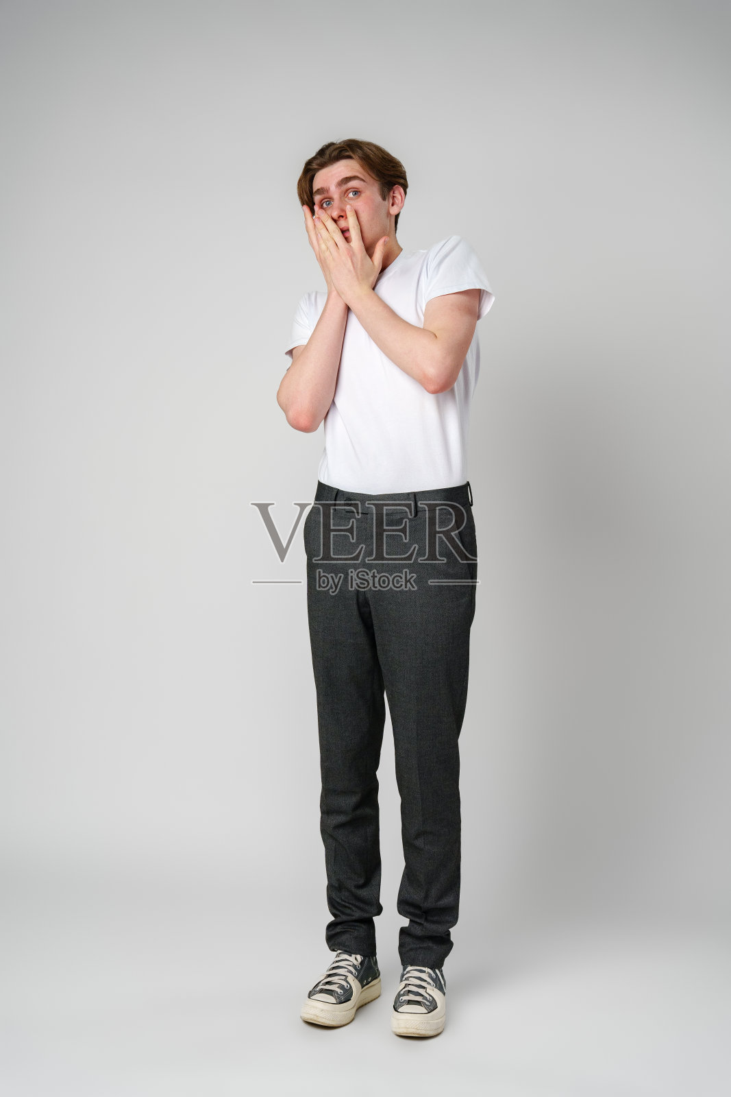 身穿白色t恤和灰色裤子的年轻人双手捂着脸,露出惊讶的表情照片摄影图片