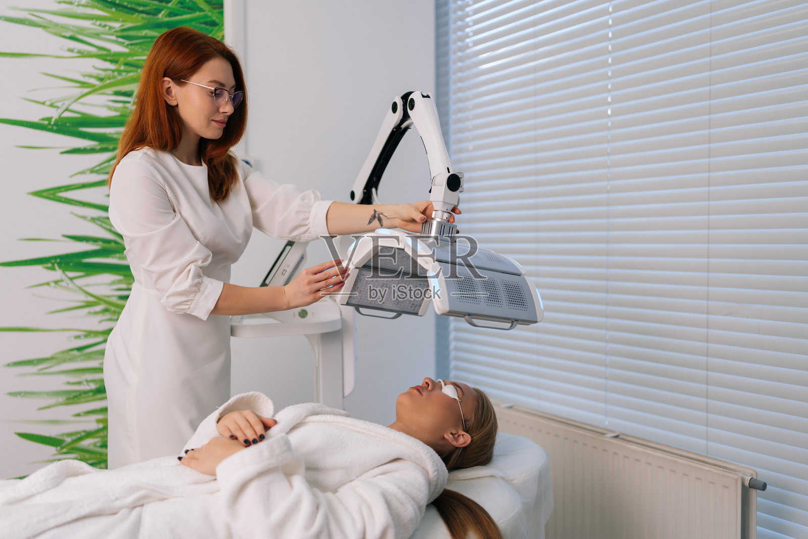 熟练的美容师专家在美容诊所连接和调整创新的皮肤嫩肤装置。一名女性患者接受蓝色LED光疗法。照片摄影图片