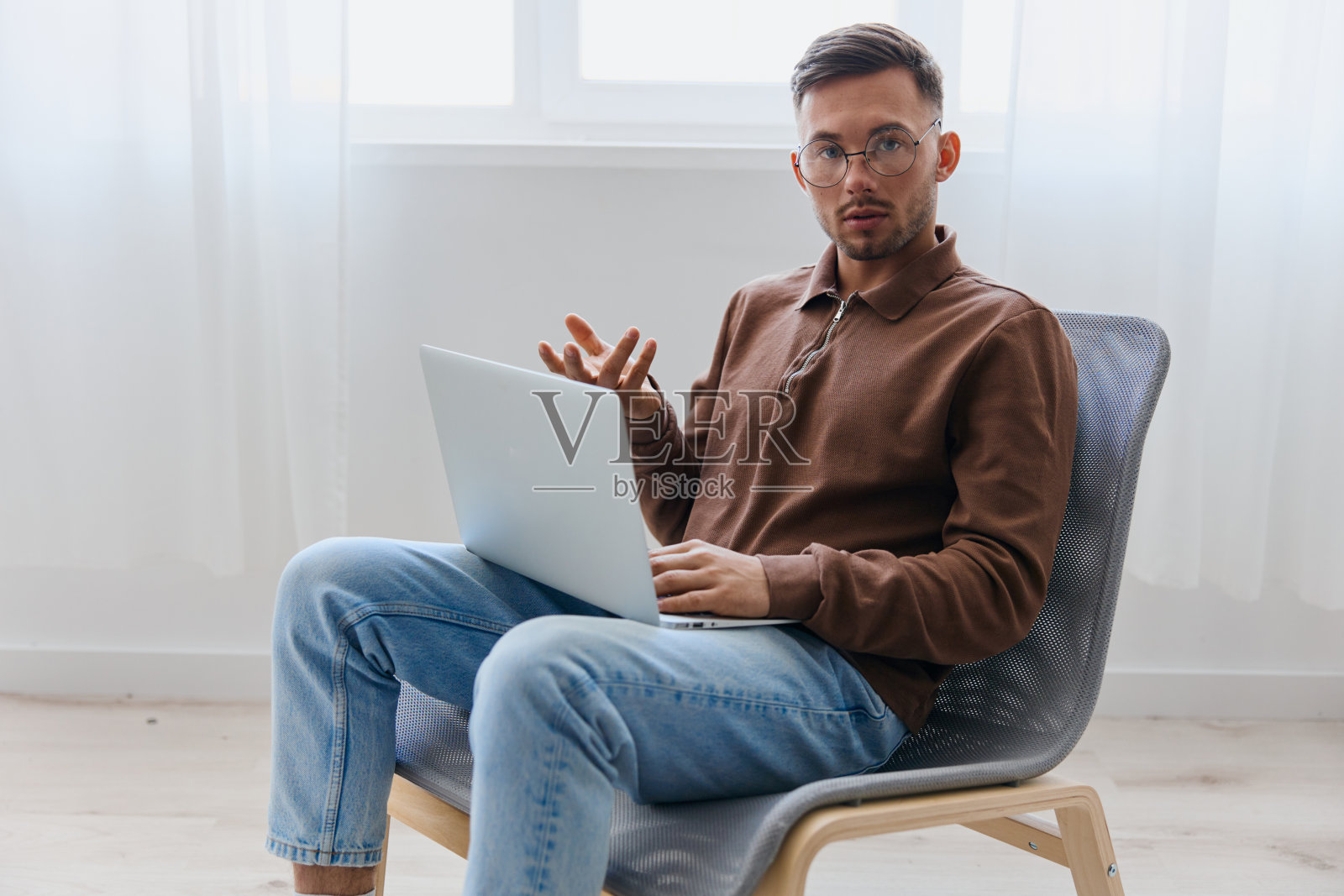 戴着眼镜的沉思严肃的年轻人举起手,坐在家里的椅子上看着相机,想着项目的最佳解决方案。远程工作远程工作沟通现代职业概念照片摄影图片