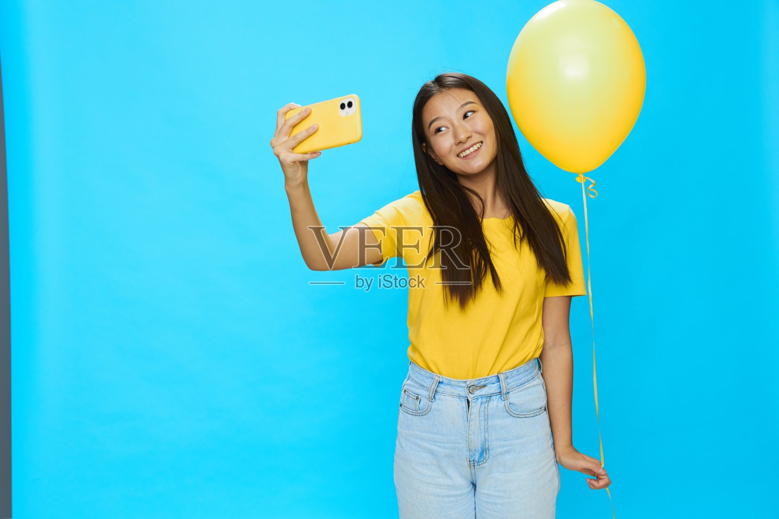 一个亚洲女人拿着手机,看着屏幕在视频通话,她手里拿着一个黄色的气球,在蓝色的背景上,穿着黄色的t恤,微笑着,露出牙齿照片摄影图片
