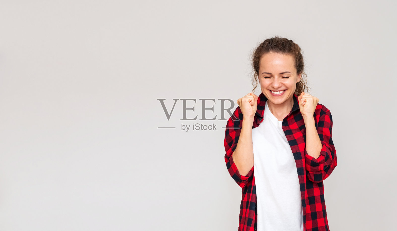 快乐的女人。幸福的表情。在画室里,一个女人闭着眼睛,握紧拳头,在平淡的背景下高兴地笑着。照片摄影图片